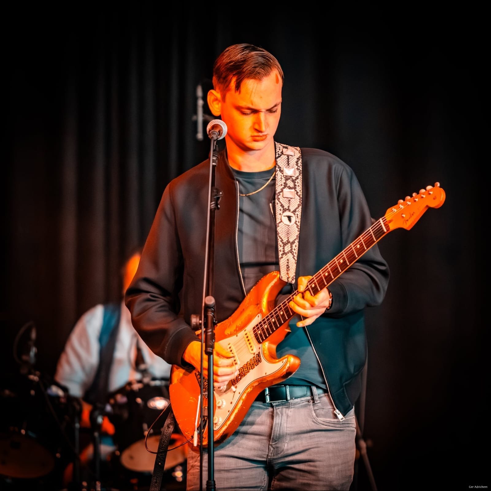 Gerbman at the microphone with sunburst relic Stratocaster, amber stage lighting, drummer in background — photo Ger Adrichem